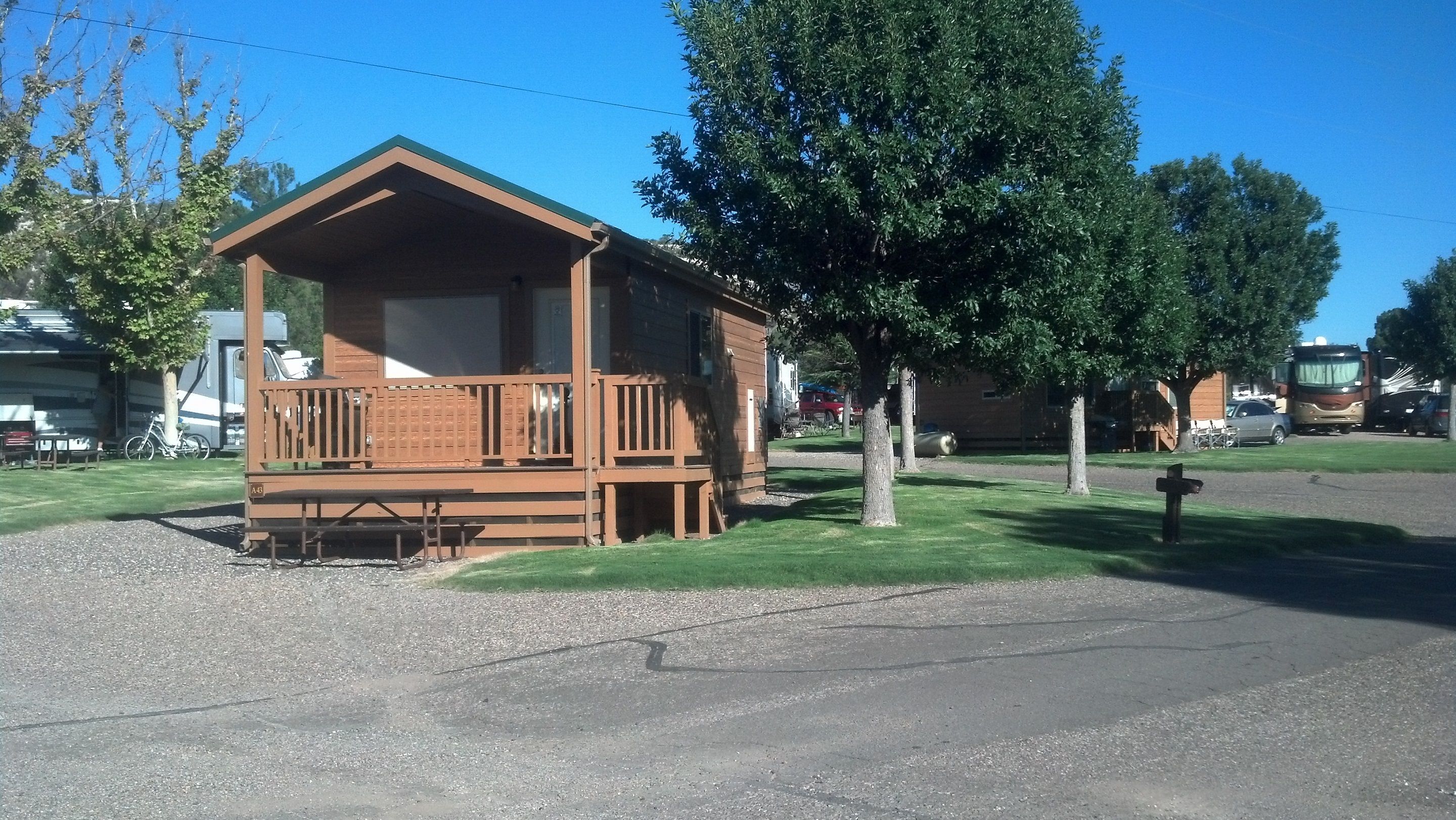 A small wooden cabin with a balcony, surrounded by trees and parked vehicles.