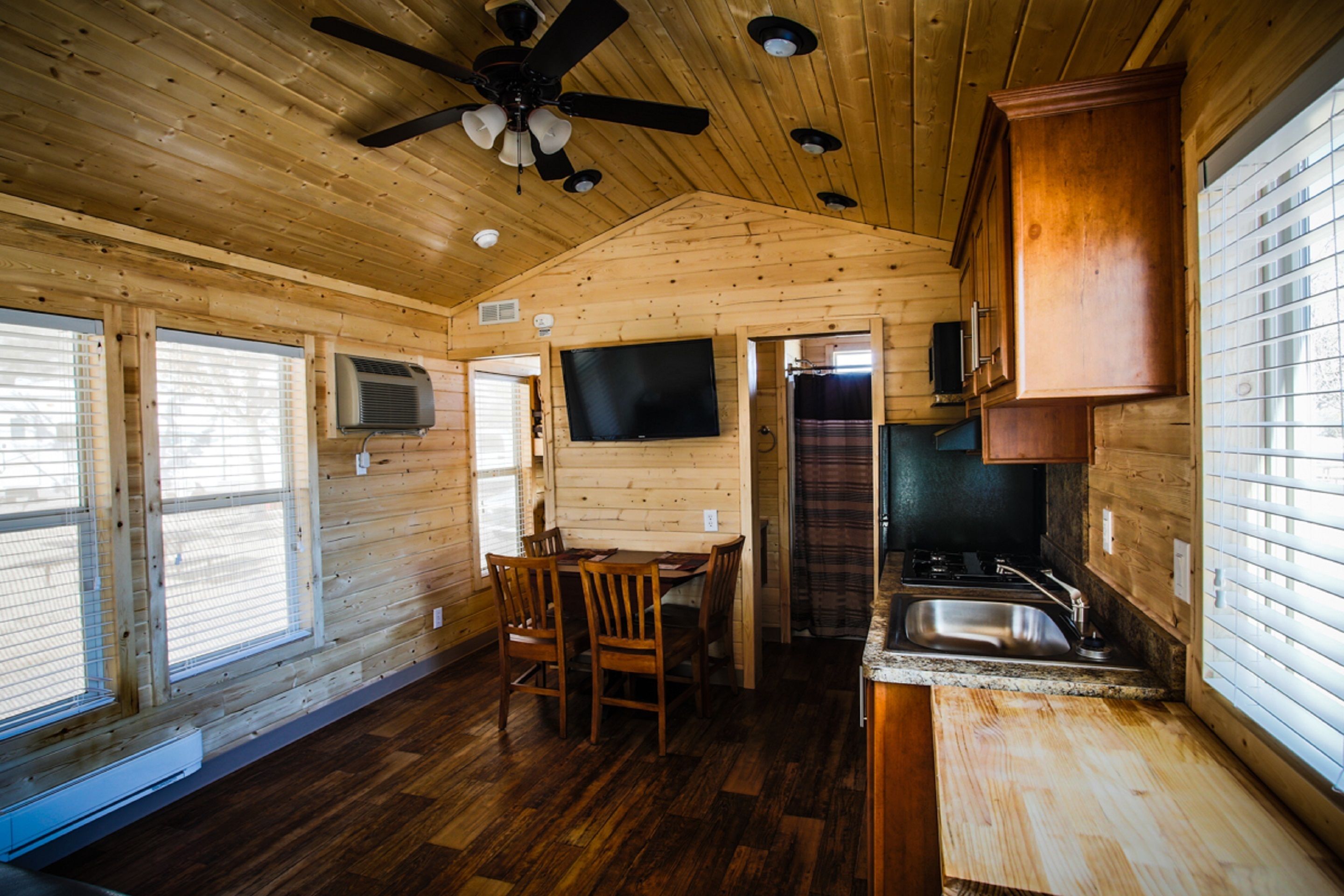 Interior de una cabaña de madera con comedor, cocina y ventilador de techo.