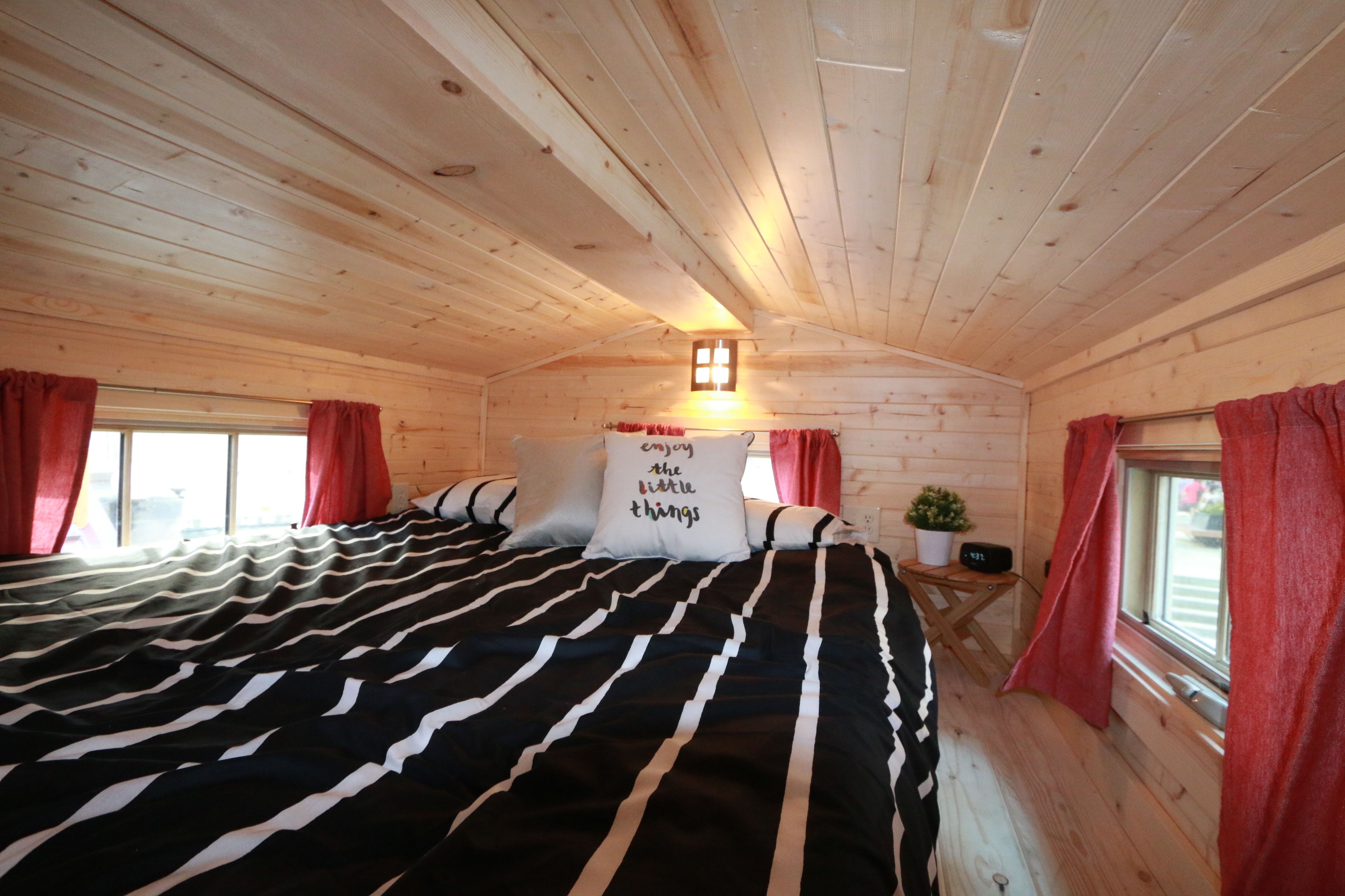 A cozy wooden cabin room with a large bed, red curtains, a small table with a plant, and a wall-mounted light fixture.