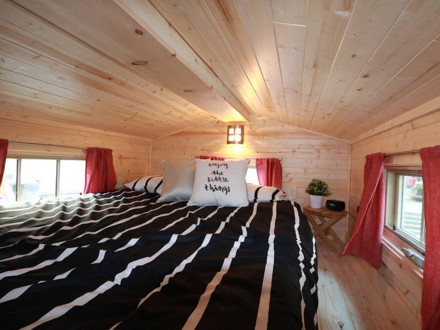 A cozy wooden cabin room with a large bed, red curtains, a small table with a plant, and a wall-mounted light fixture.