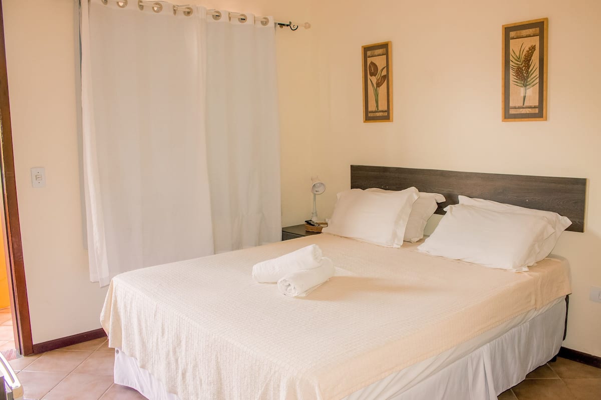 A neatly made bed with white linens and pillows, a wooden headboard, and two framed botanical prints on the wall.