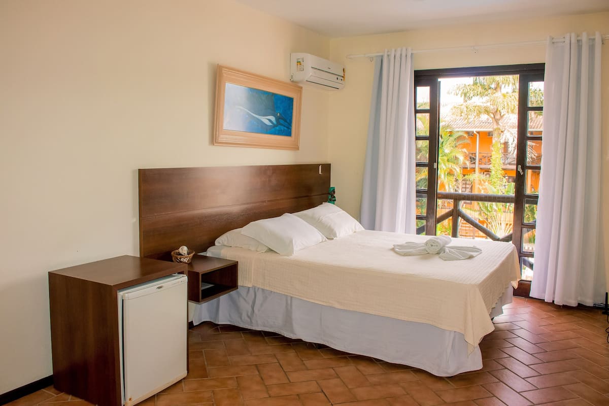 A hotel room with a wooden headboard bed, a small refrigerator, a painting on the wall, and a view of the outside through a window with curtains.