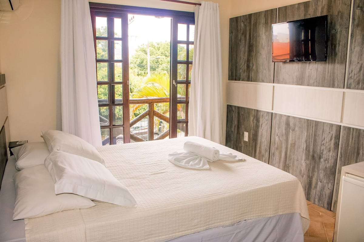 A hotel room with a bed, a TV mounted on the wall, and a balcony with a view of greenery.