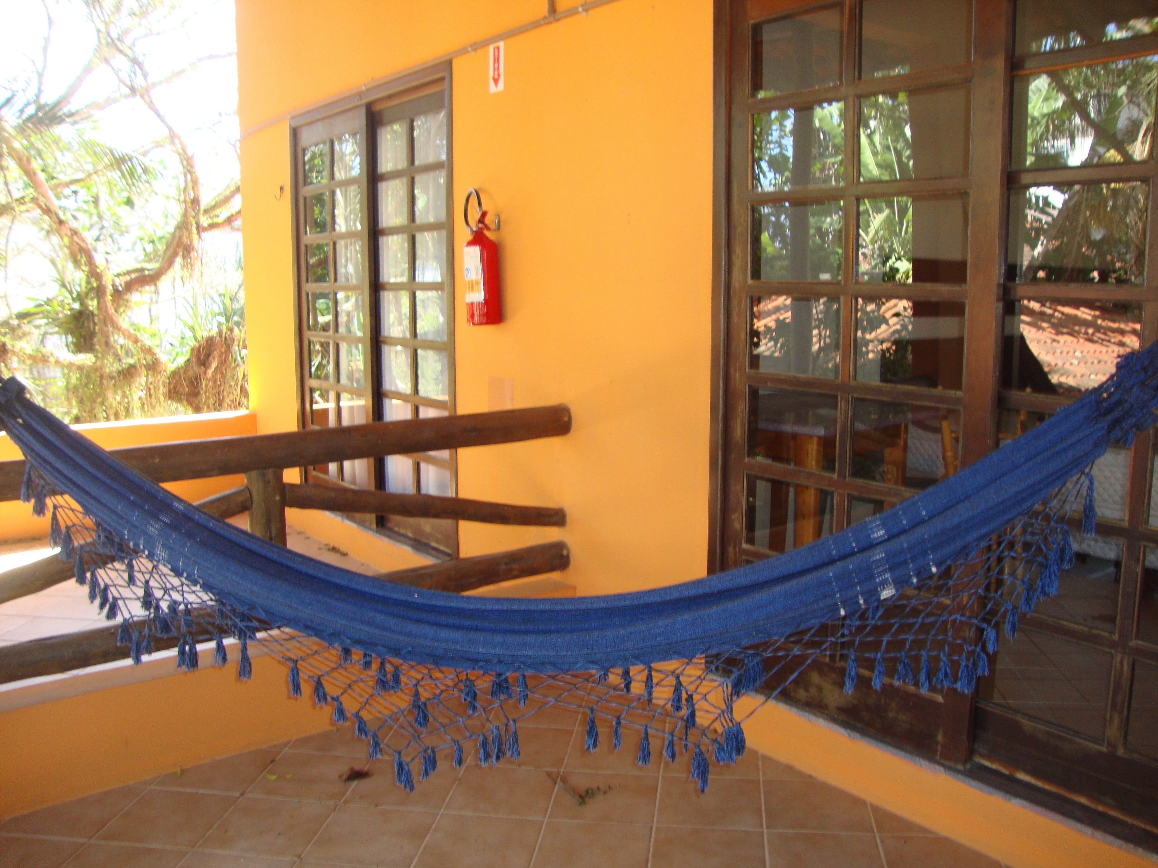 A hammock on a balcony with a fire extinguisher on the wall and a window with a view of trees.