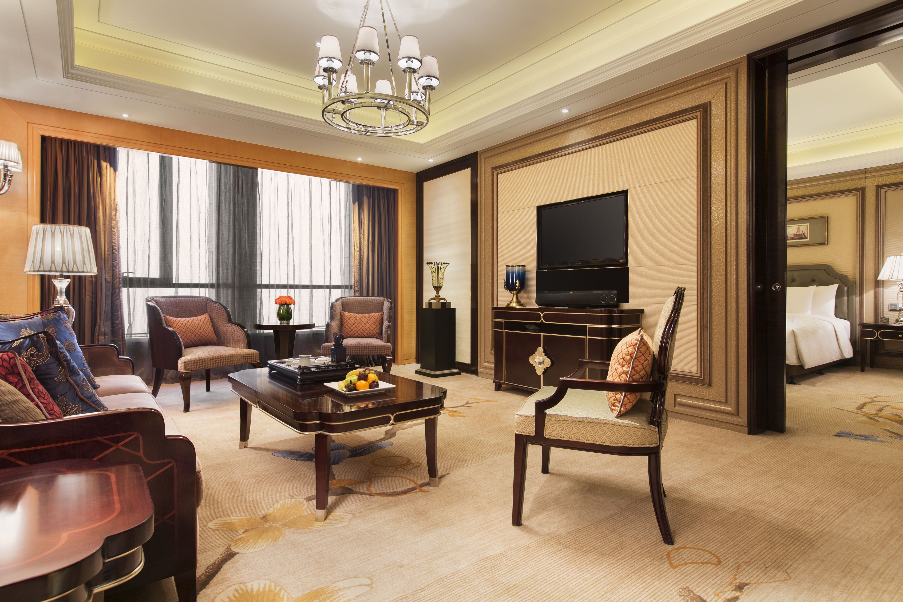 A spacious hotel room with a large flat-screen TV, a dining area with a coffee table, and a seating area with two armchairs and a sofa.