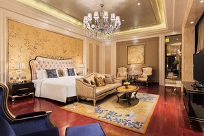 A luxurious hotel room with a large bed, a sofa, two armchairs, a coffee table, and a TV.