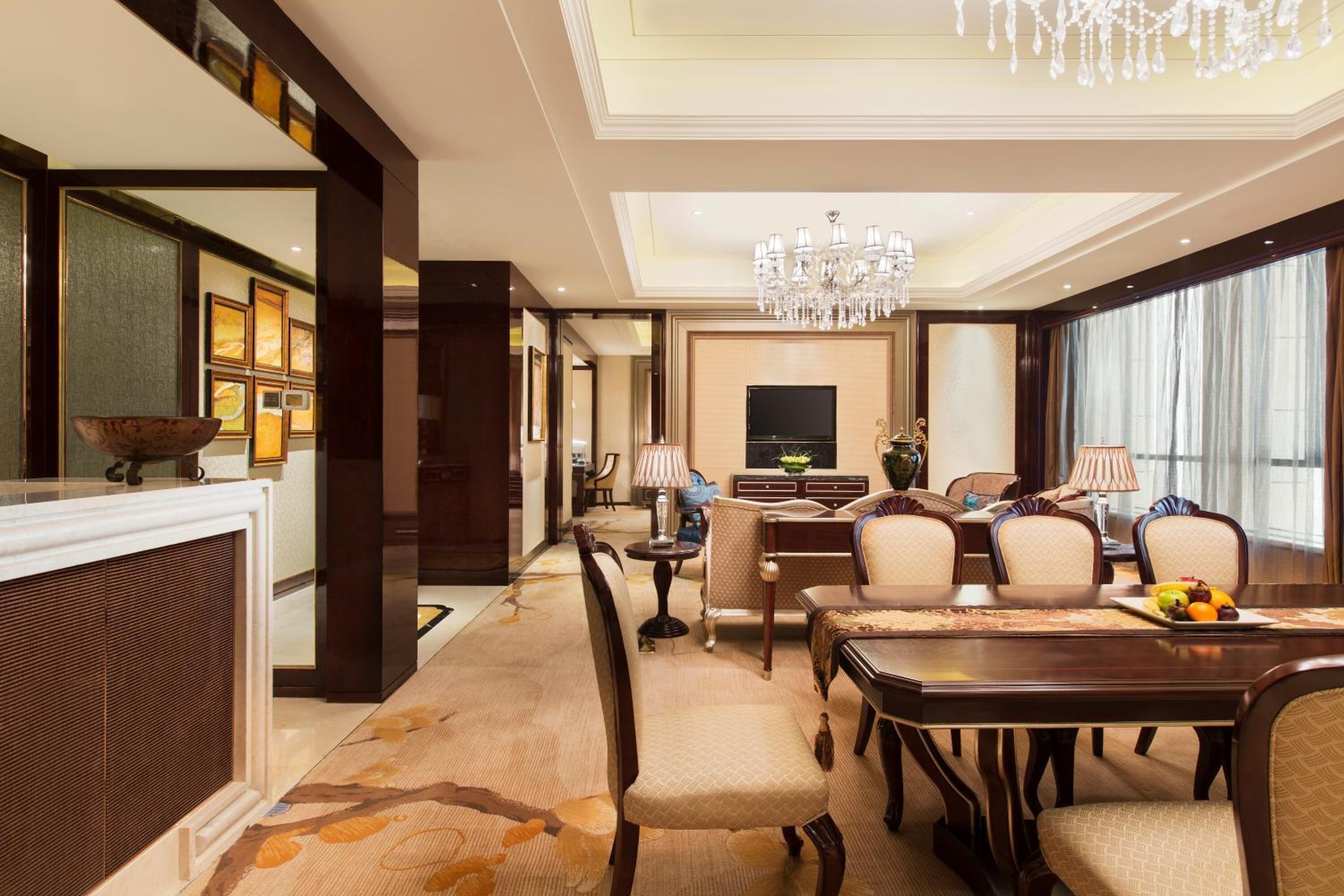 Un hallo de hotel espacioso con una gran mesa de comedor, asientos cómodos, un candelabro y un televisor.