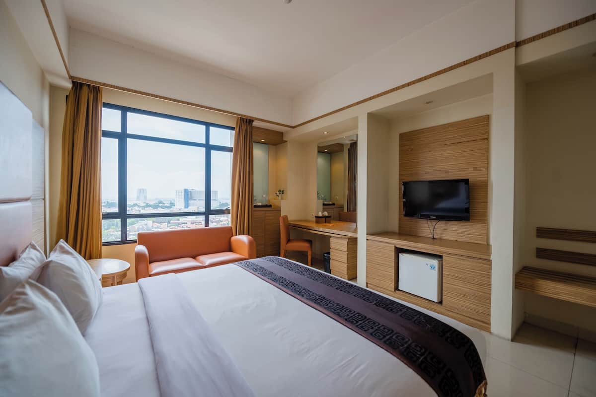 A hotel room with a large bed, a desk, a television, and a view of the city through large windows.