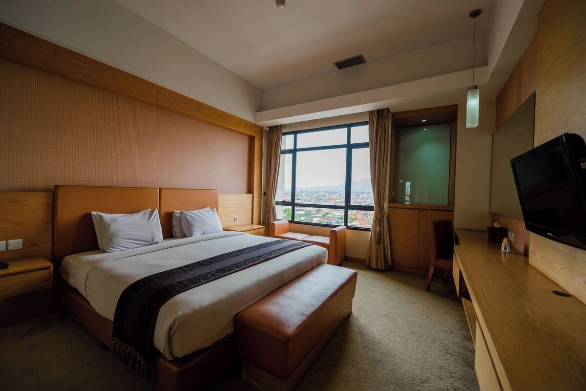 A hotel room with a large bed, a wooden desk, a television, and a window with a city view.