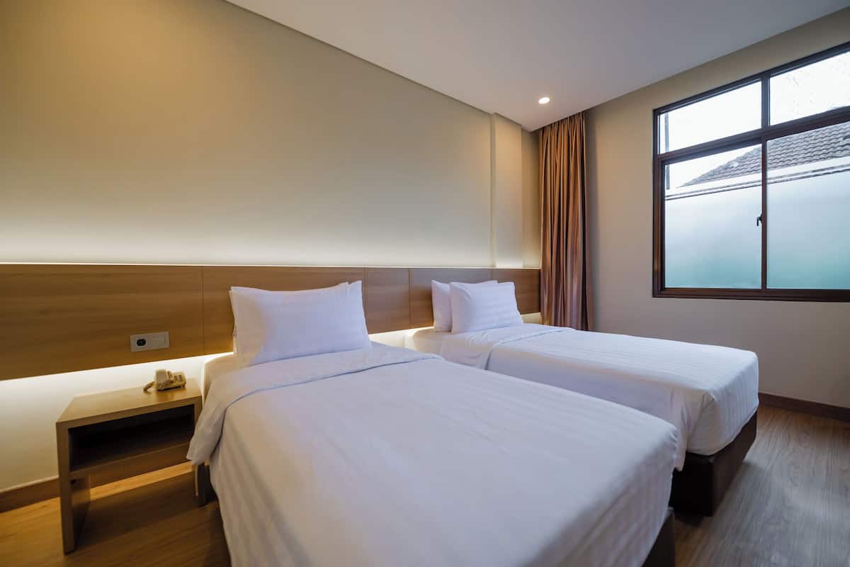 A hotel room with two beds, a bedside table with a phone, a large window with curtains, and a wooden headboard.