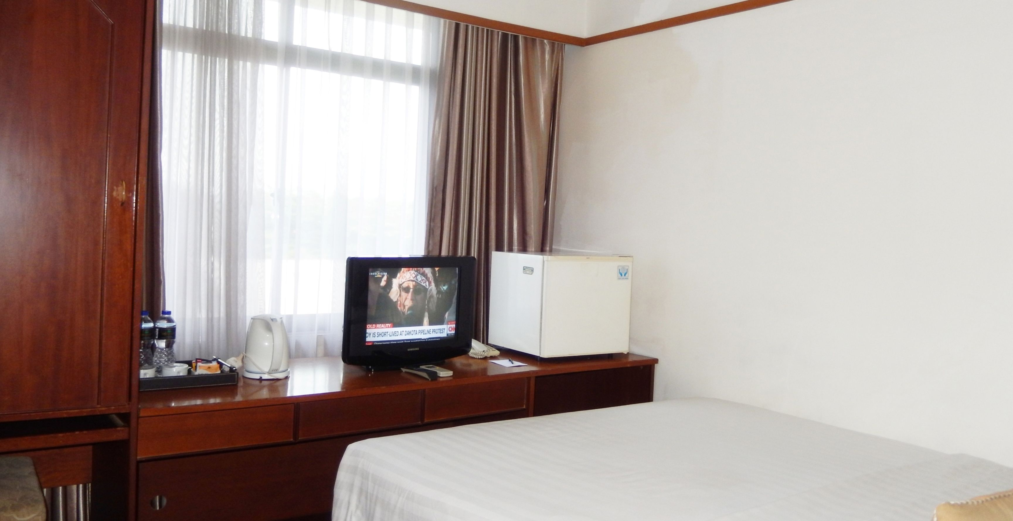 A hotel room with a bed, a television on a wooden dresser, a mini-fridge, and a window with curtains.