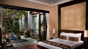 A hotel room with a large bed, a bedside table, a lamp, and a view of a garden through an open door.