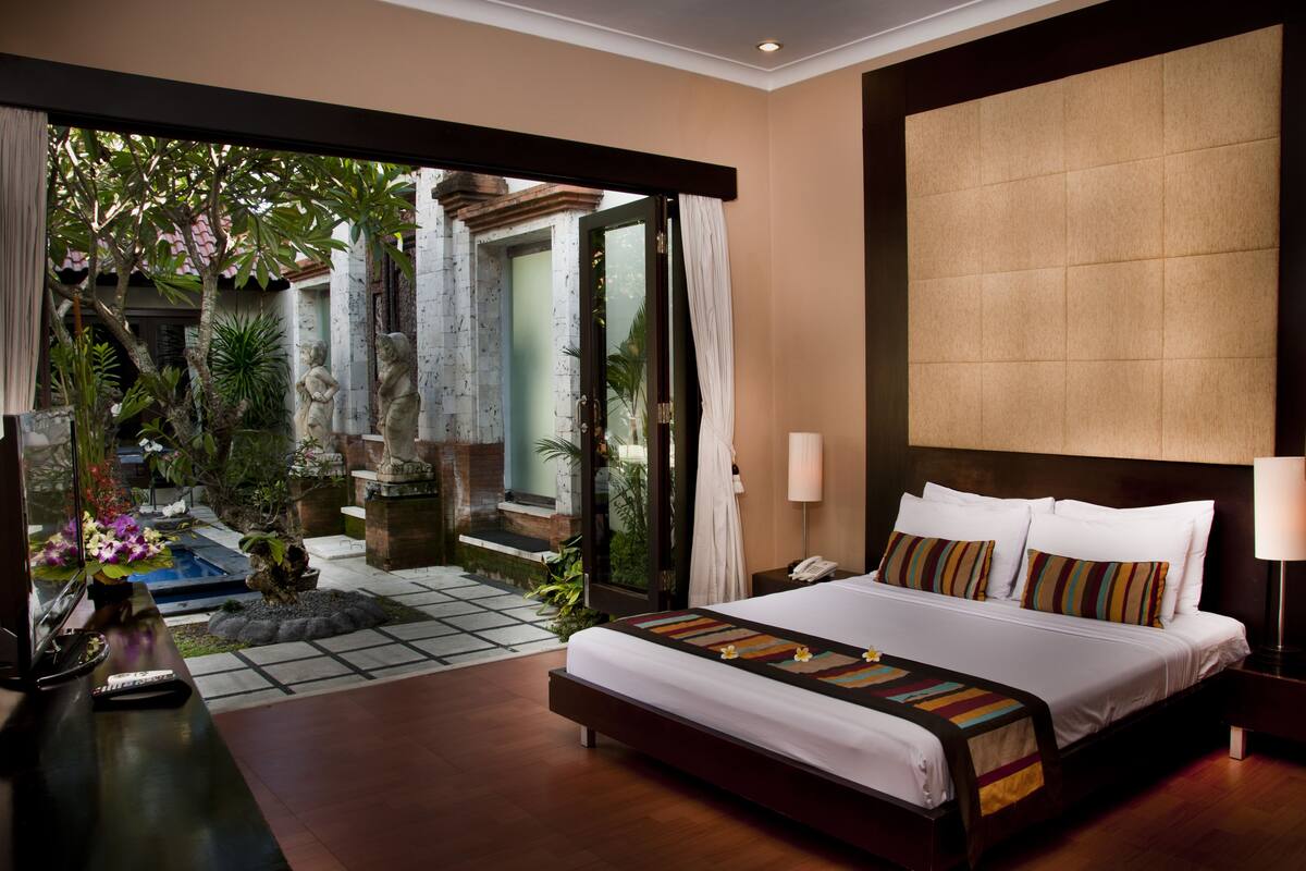 A hotel room with a large bed, a bedside table, a lamp, and a view of a garden through an open door.