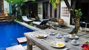 A poolside dining area with a wooden table set for breakfast, surrounded by lounge chairs and a small garden.