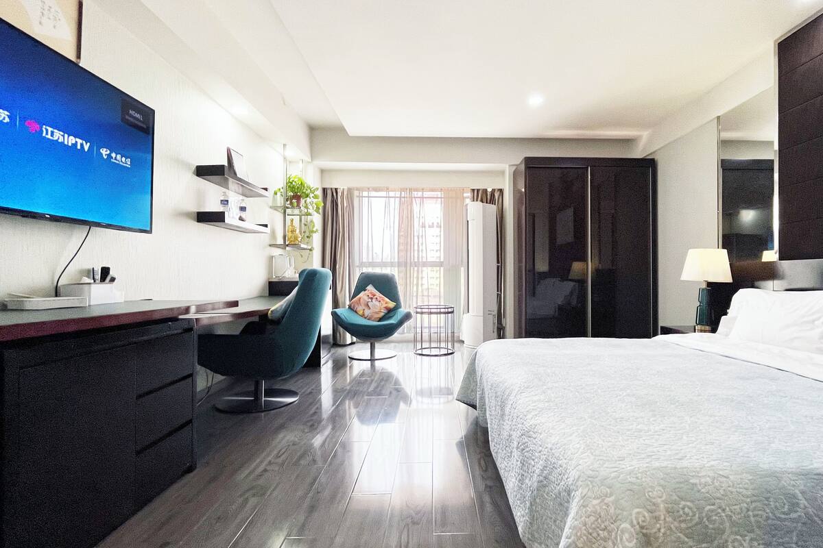 A hotel room with a bed, a desk with a TV, a chair, a small table, and a wardrobe.