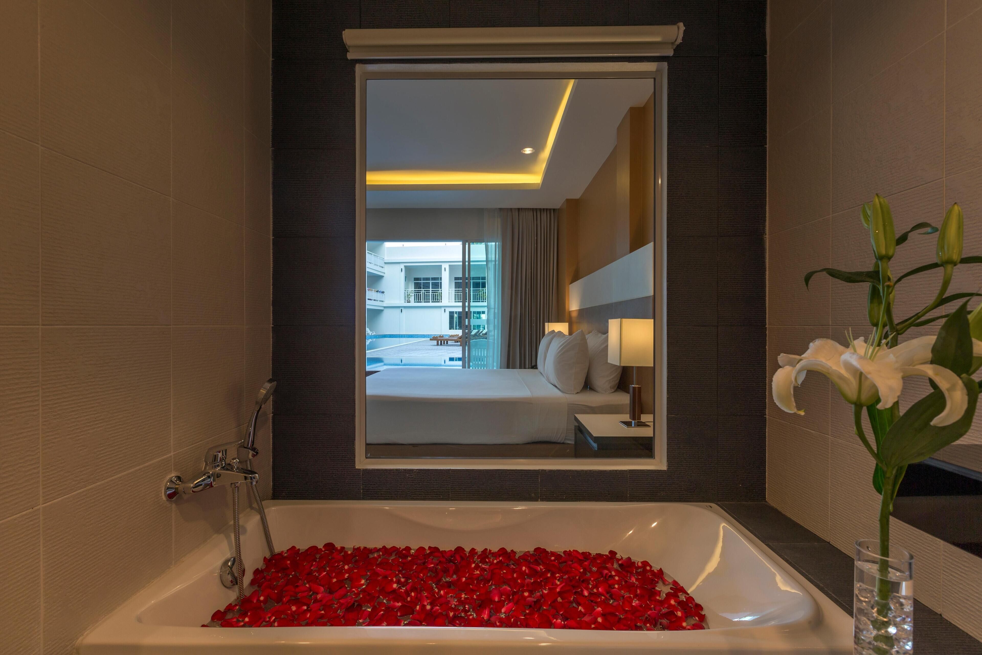 Une salle de bain avec une baignoire remplie de pétales de roses, un miroir reflétant une chambre d’hôtel et un vase avec des fleurs blanches.
