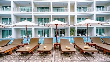 A hotel pool area with multiple lounge chairs and umbrellas, a tiled floor, and a pool with clear blue water.