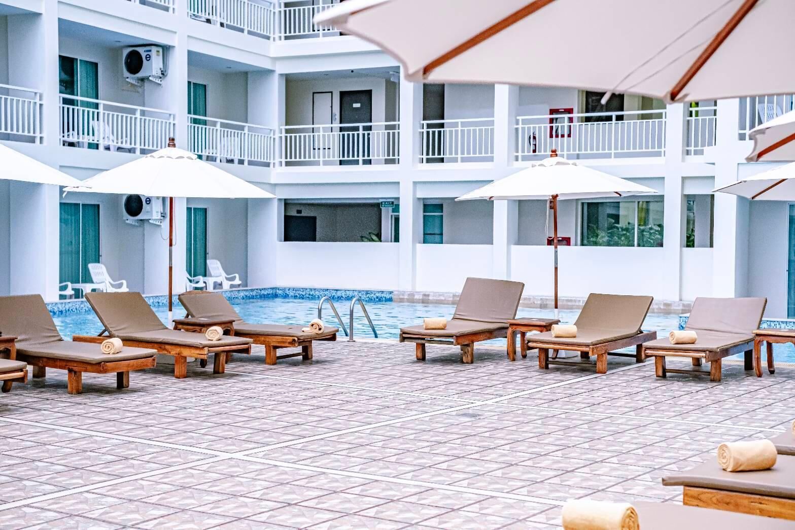 Un espace aménagé au bord de la piscine, avec des chaises longues et des parasols, un sol carrelé et un bâtiment doté de nombreux balcons.