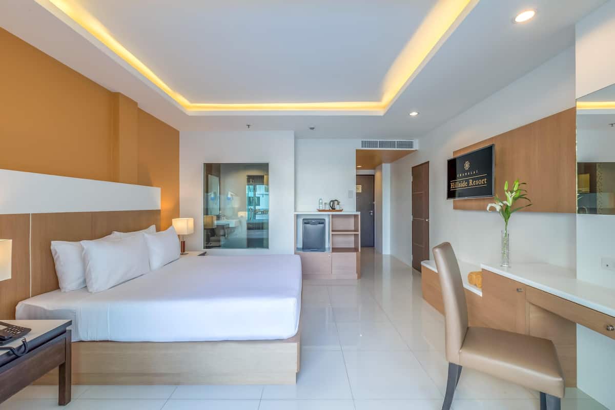 Une chambre d’hôtel moderne avec un grand lit, un bureau et une chaise. On aperçoit un petit coin cuisine au fond de la pièce.