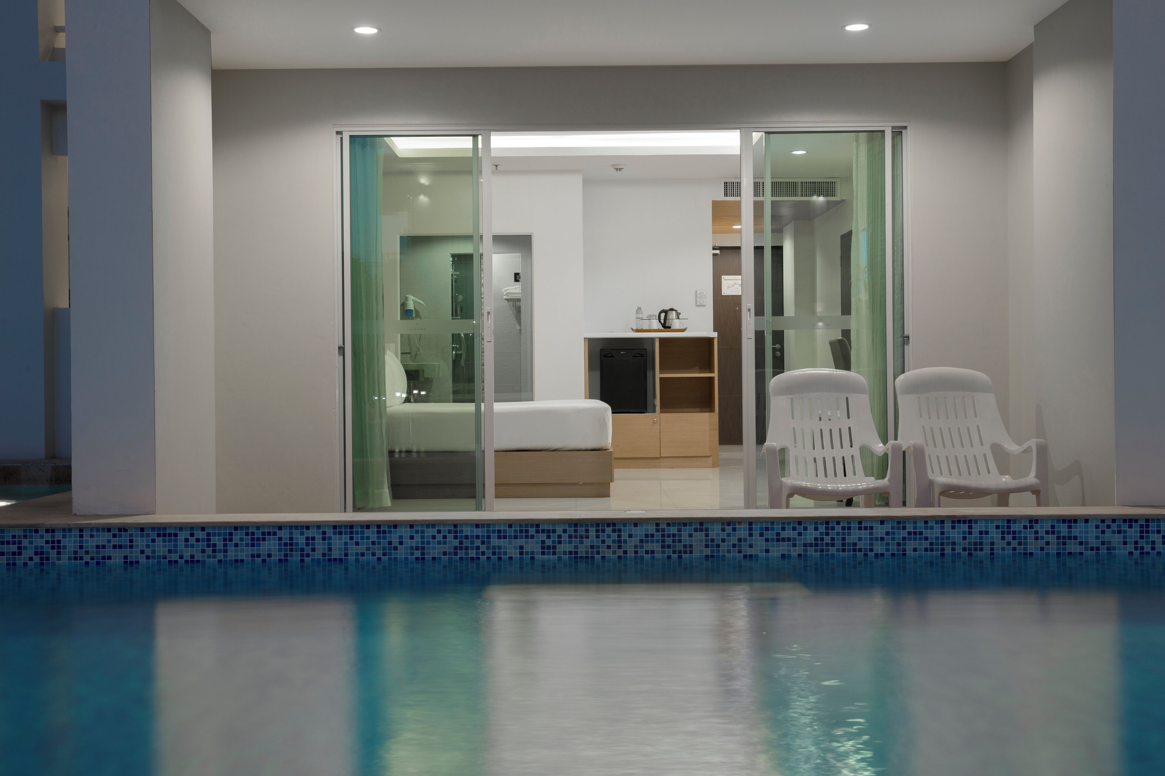 A modern hotel room with a glass door leading to a bathroom, a bed, a small kitchen area, and two white chairs by a pool.