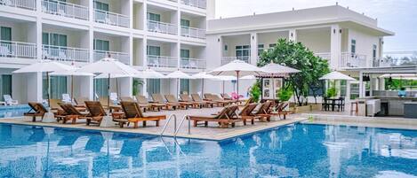 A swimming pool with lounge chairs and umbrellas, surrounded by a multi-story building with balconies.