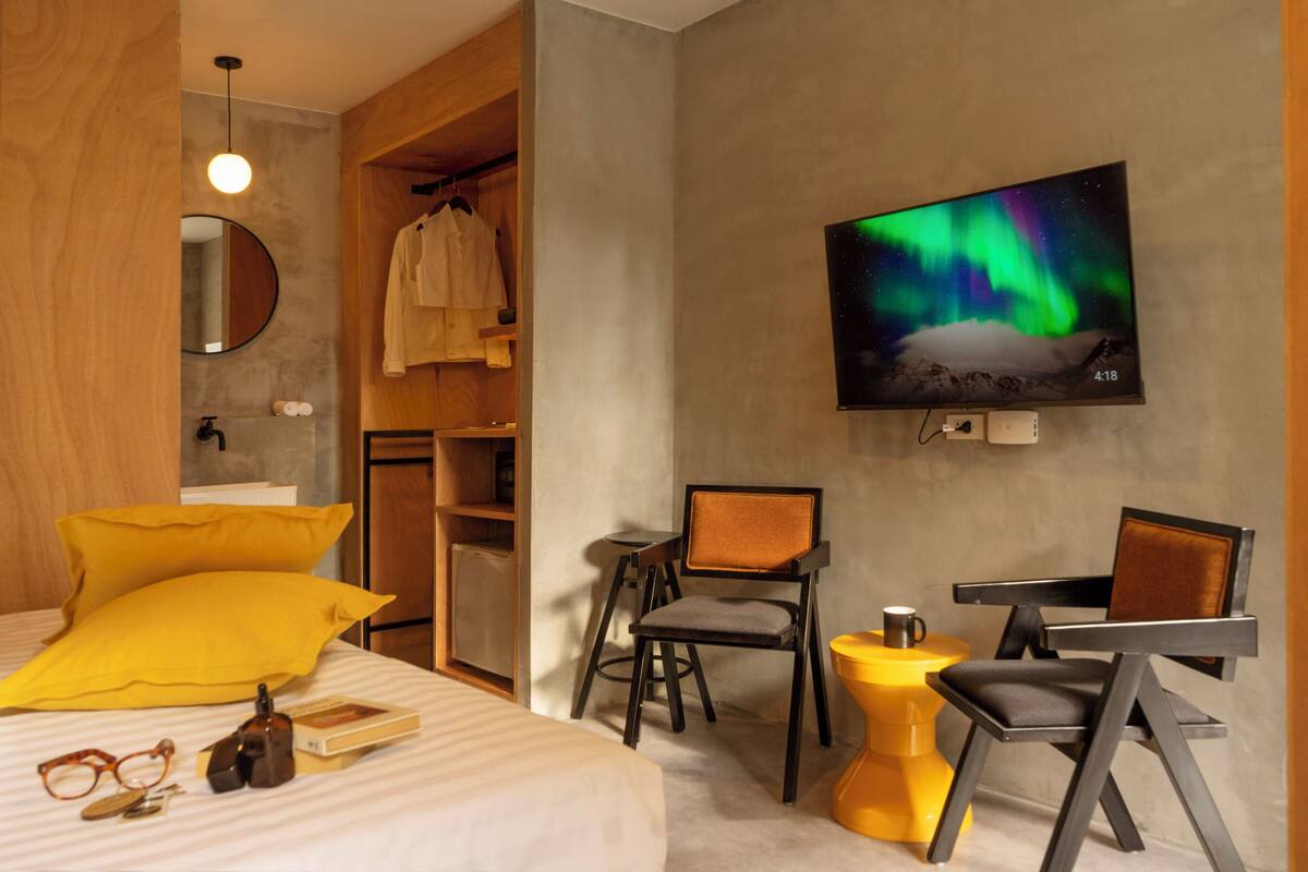 A modern hotel room with a bed, a TV displaying the Northern Lights, a wooden wardrobe, and a small seating area with chairs and a side table.