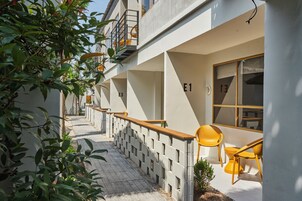 A modern building with numbered rooms E1, E3, and E13, a wooden balcony, and yellow chairs outside.
