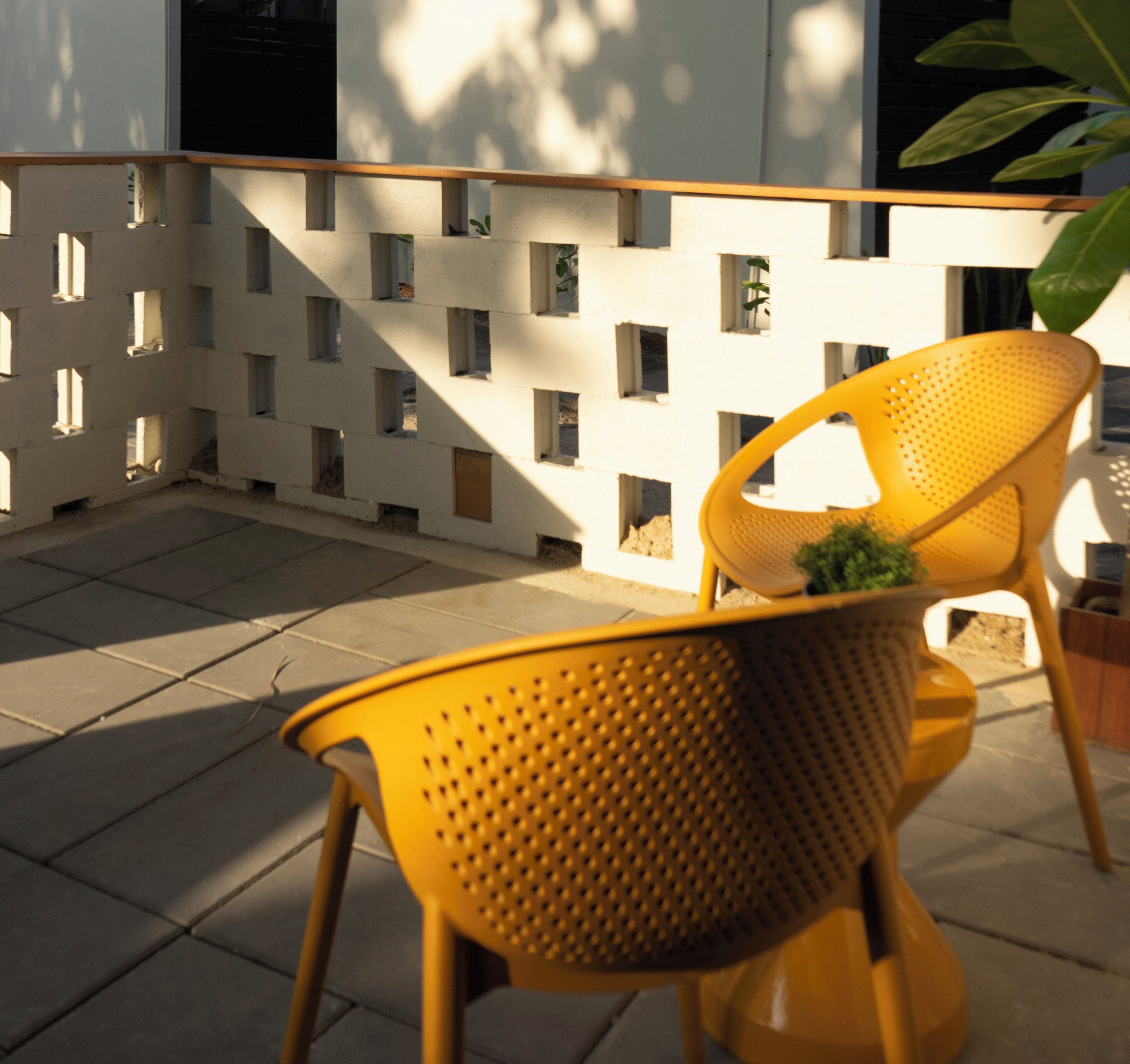 A yellow chair with a perforated backrest and a small plant on it, positioned on a tiled patio area with a white wall featuring square cutouts in the background.