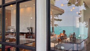 A restaurant with a view of the sea, featuring a dining area with tables and chairs, and a kitchen visible through the glass wall. - Escape Beach Resort (Koh Samui)