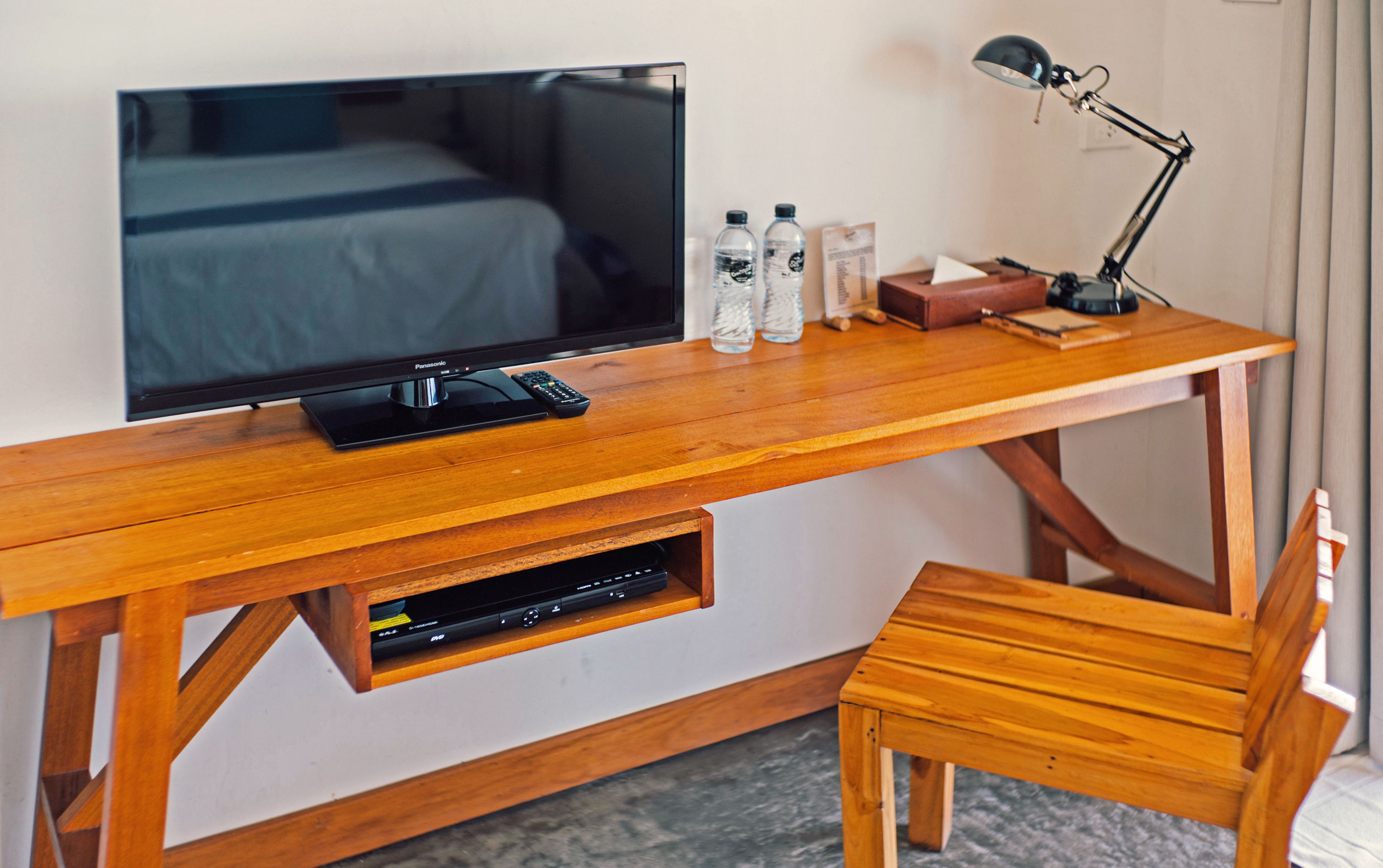 A wooden desk with a television, remote control, water bottles, a lamp, and a small wooden chair.