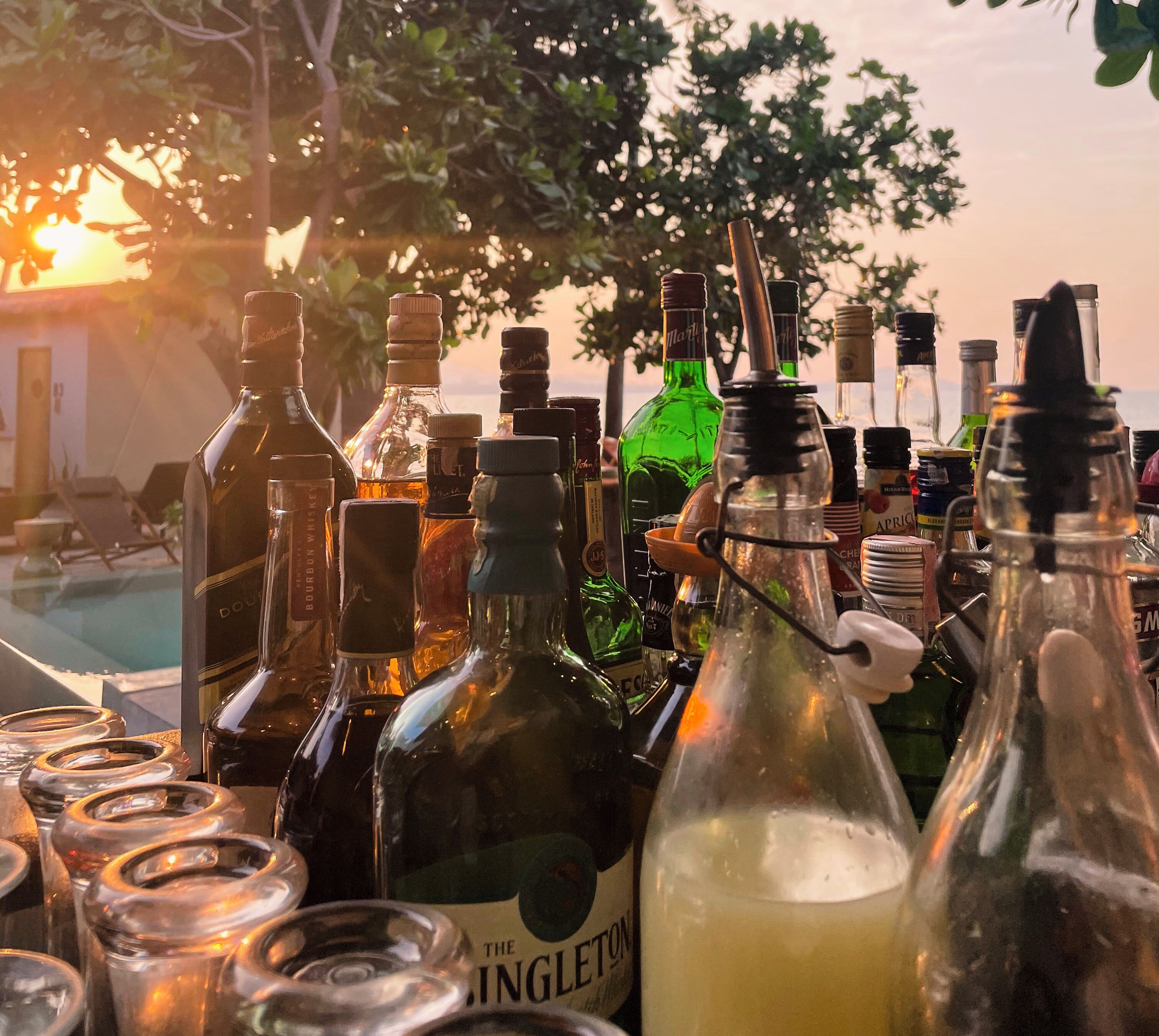 A variety of bottles and glasses on a table, likely for serving drinks, with a sunset in the background.