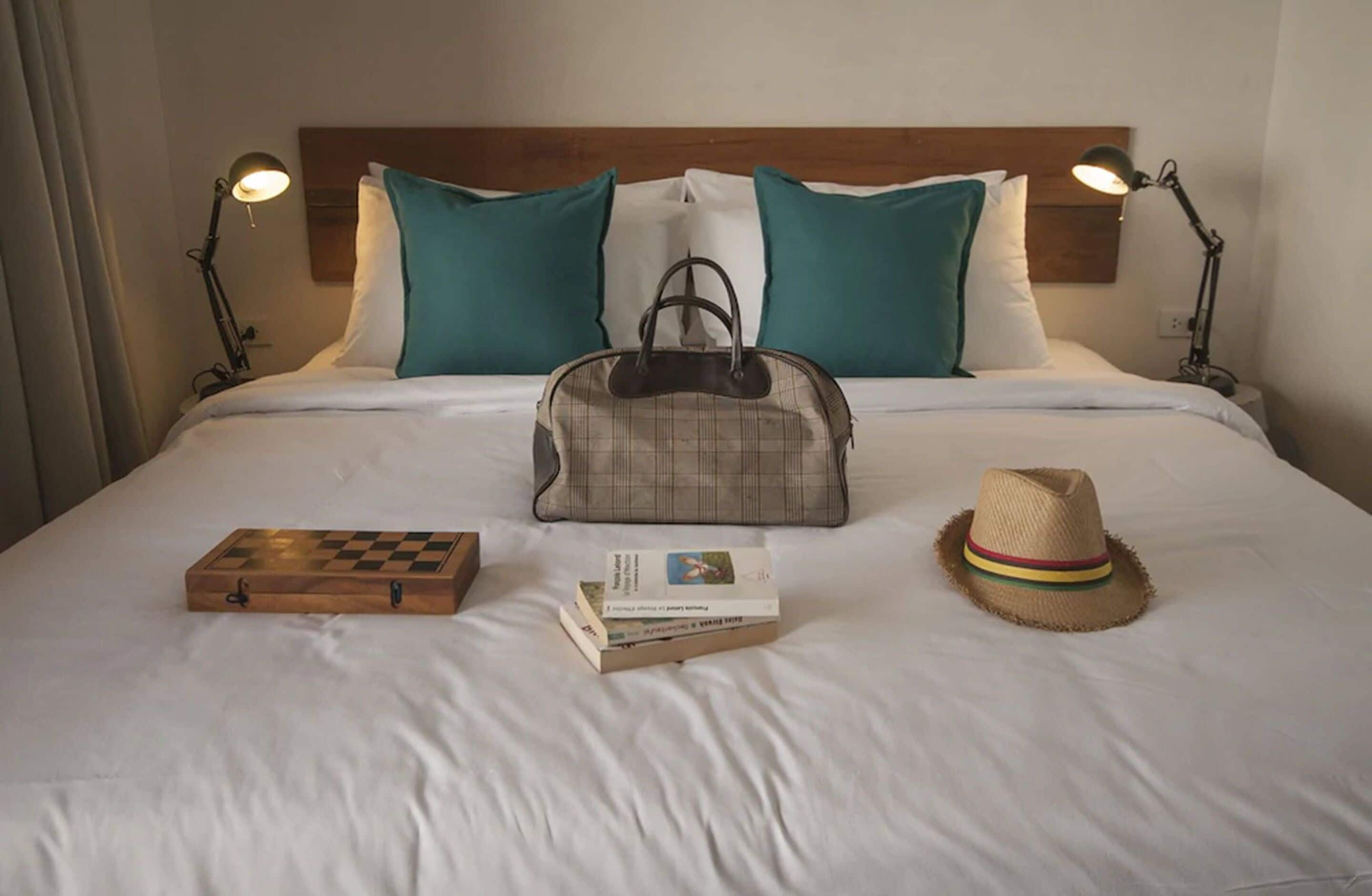 Una cama bien hecha con cabecera de madera, dos almohadas color turquesa, un bolso de cuadros, un tablero de ajedrez, una pila de libros y un sombrero de paja.