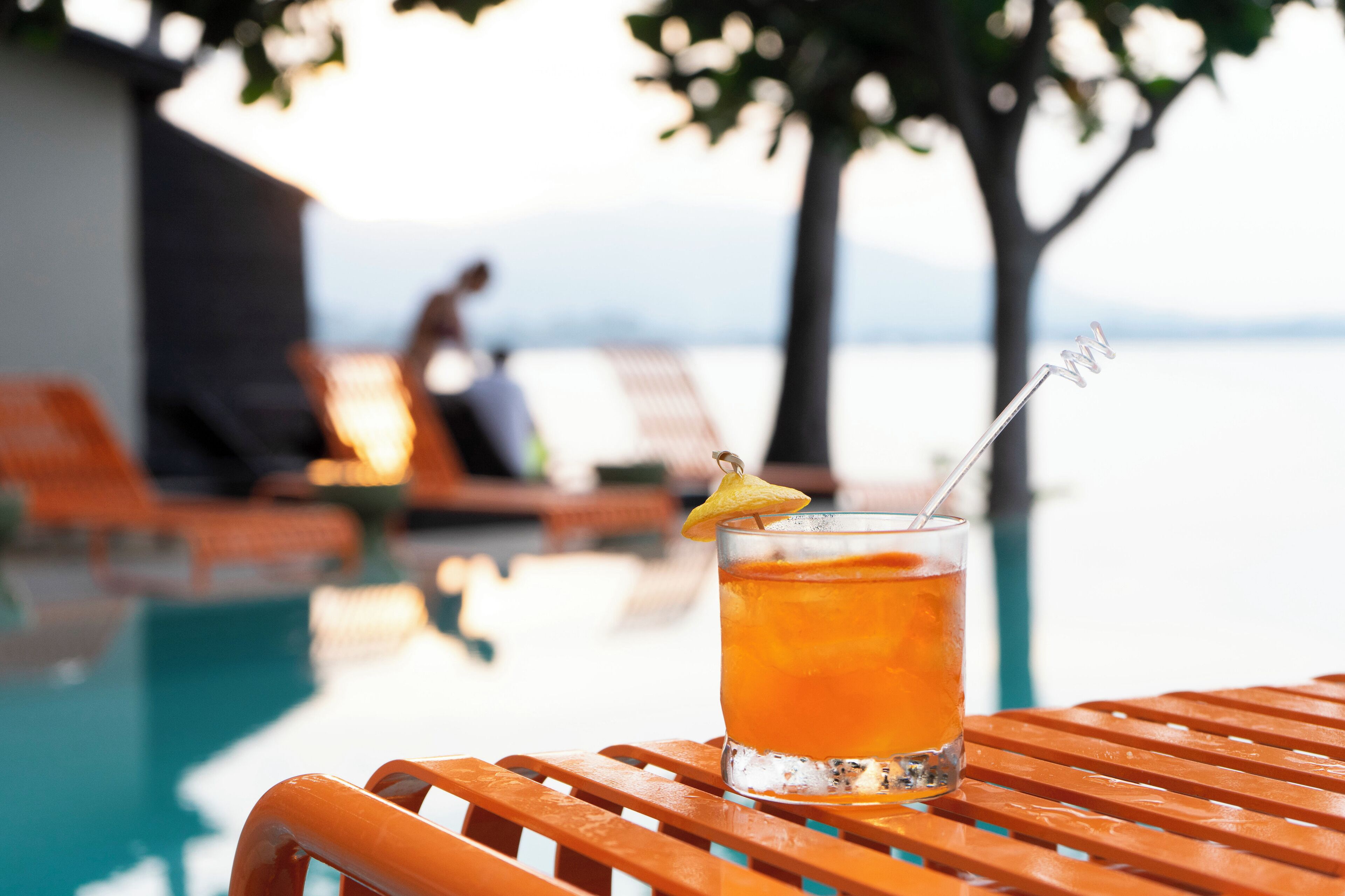 Un vaso de bebida color ámbar con una rodaja de limón sobre una mesa de madera junto a una piscina.