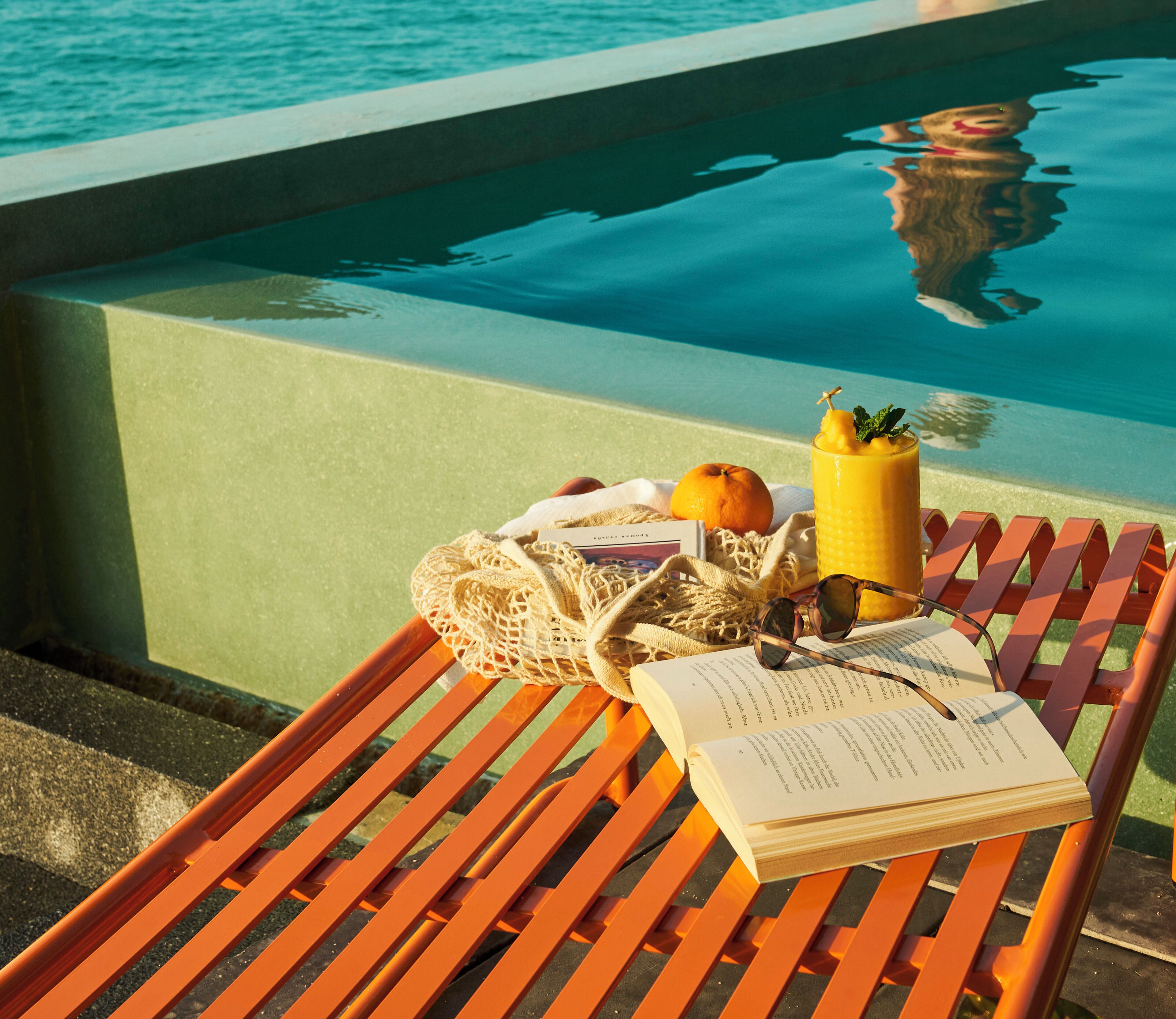 Una silla de lounge de madera junto a una piscina, con un libro, una naranja y una bebida.