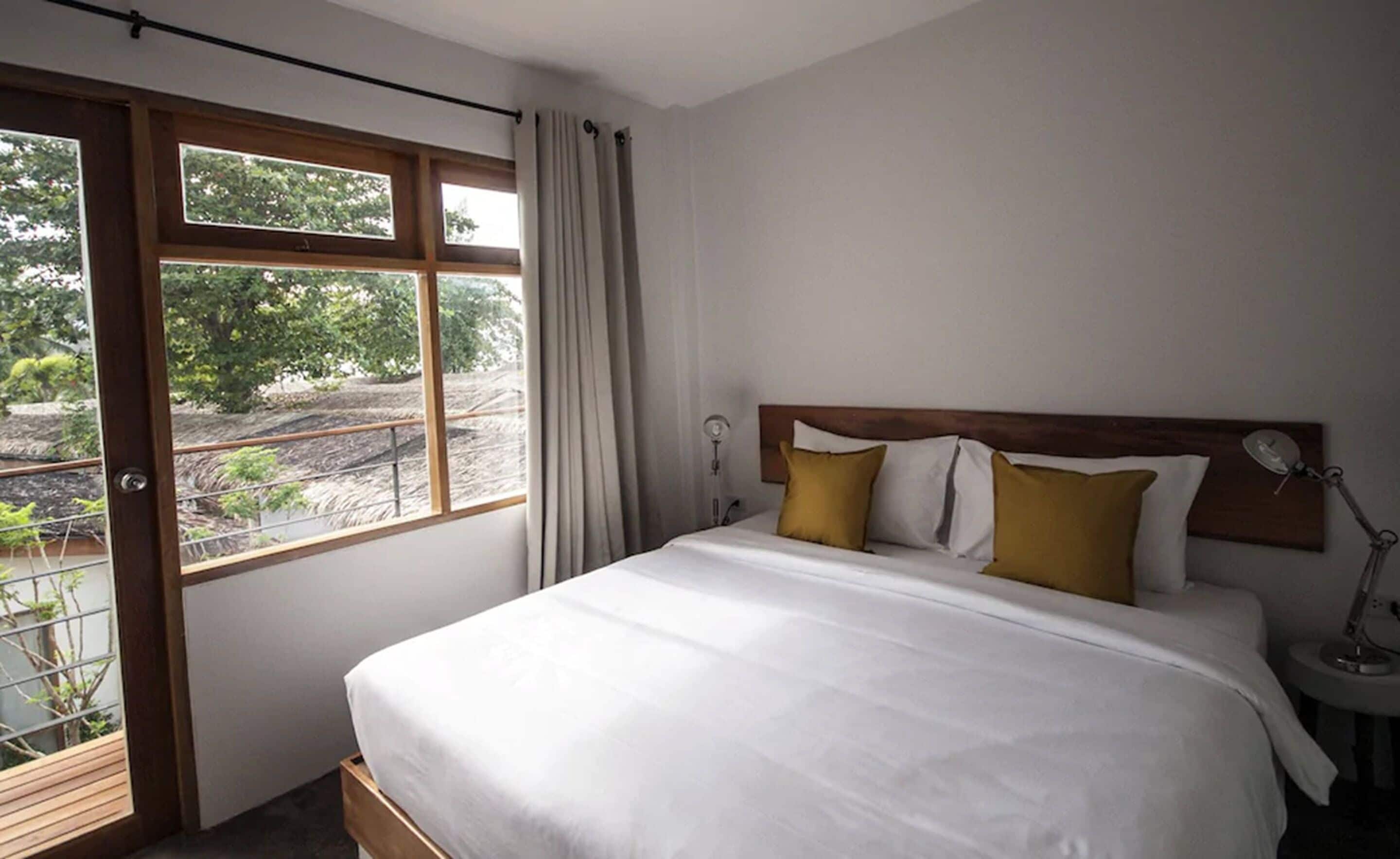 A bedroom with a large bed, a window with curtains, a bedside lamp, and a view of greenery outside.