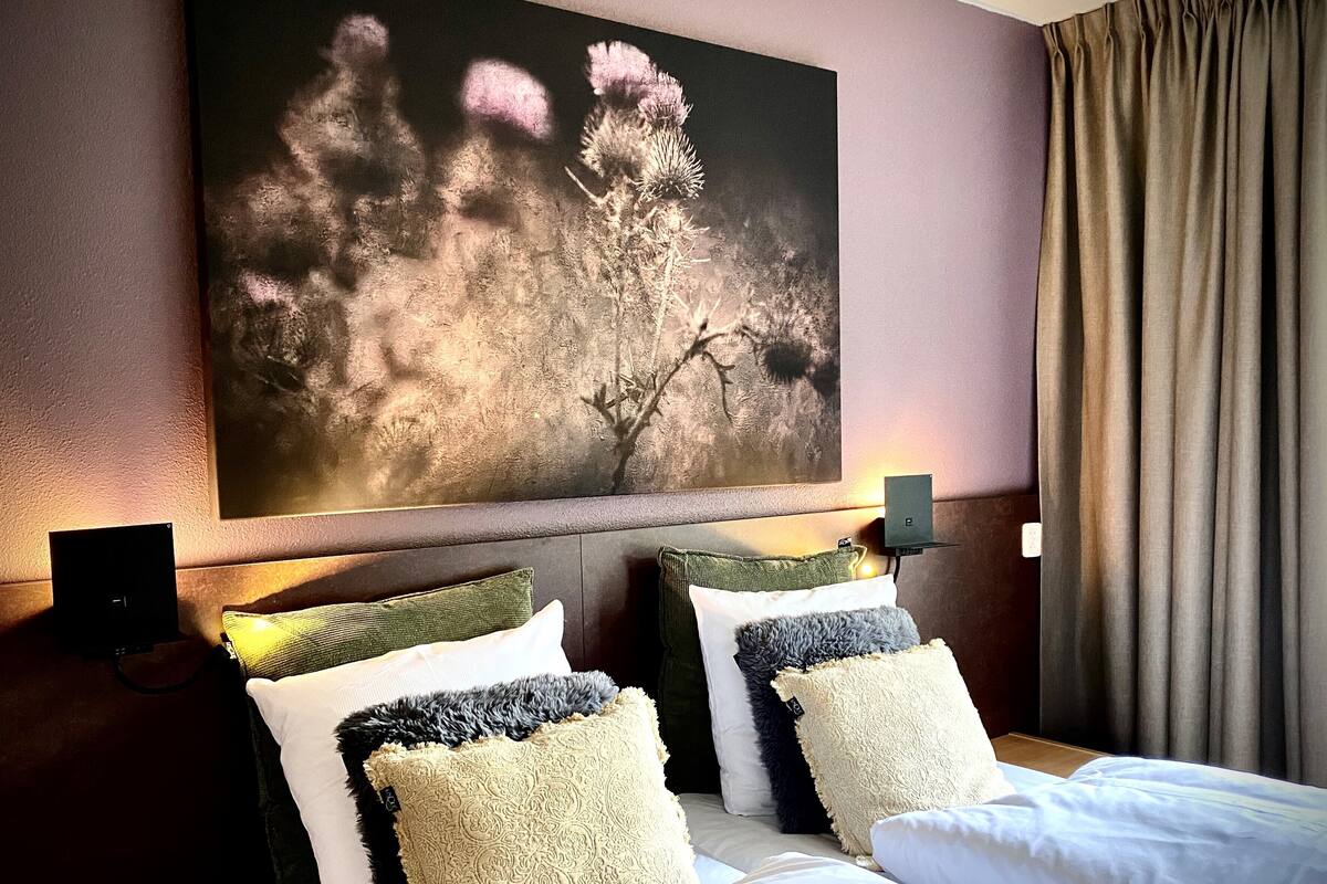 Quarto de hotel com uma grande pintura floral na parede, cama com várias almofadas e dois alto-falantes fixados na parede.