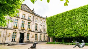 A building with classical architecture, a well-maintained hedge, and a statue of a reclining figure.