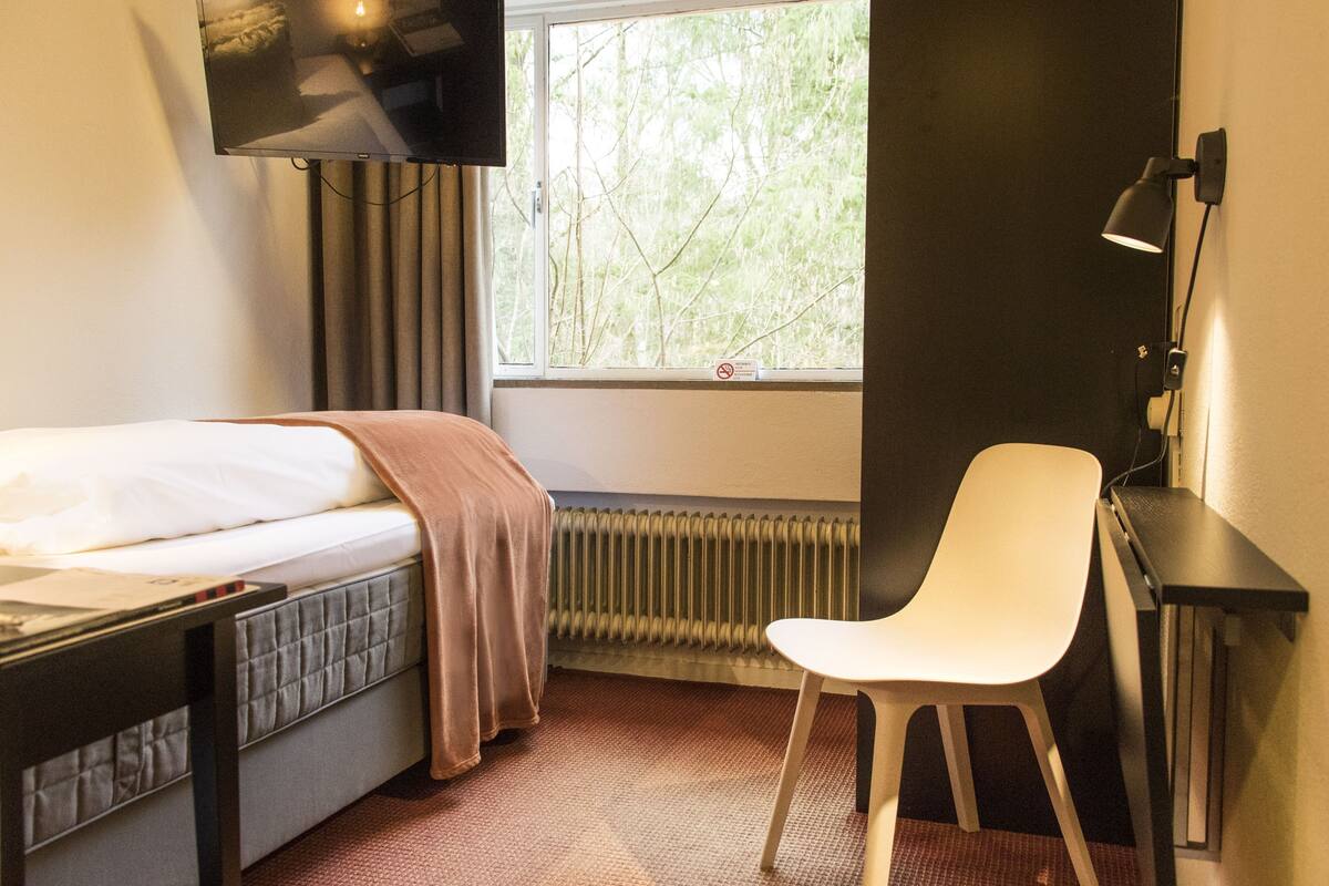 Quarto de hotel com cama, escrivaninha, cadeira, televisão fixada na parede e janela com vista para árvores.