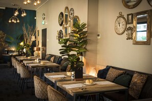 Un restaurante con varias mesas y sillas, una gran planta como adorno central, y diversas decoraciones en las paredes, incluyendo relojes y espejos.
