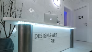 Una recepción con el rótulo "DESIGN & ART PIÉ", una laptop encima, una planta en maceta y una puerta con el número 101.