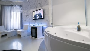 Un baño moderno con una bañera grande, un televisor montado en la pared y un rincón de estar con una mesita y una lámpara.