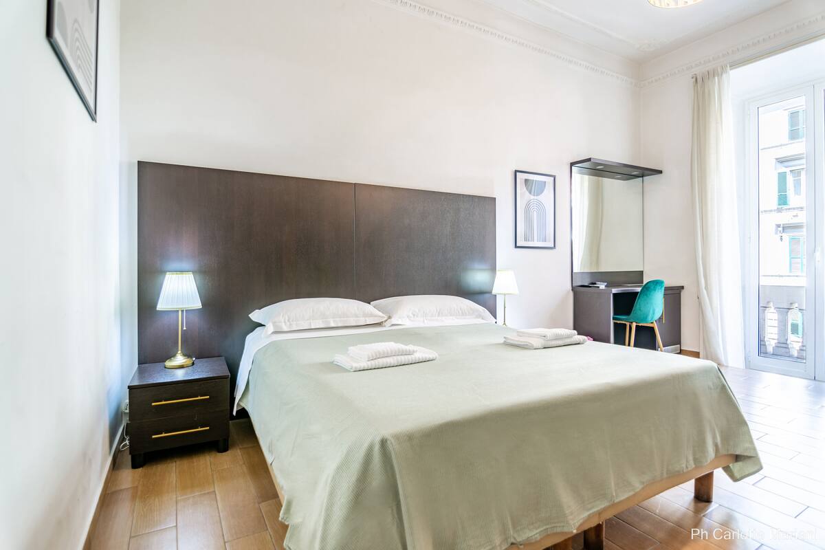 A neatly made bed with a green bedspread, a bedside lamp, a small wooden nightstand, and a desk with a chair.