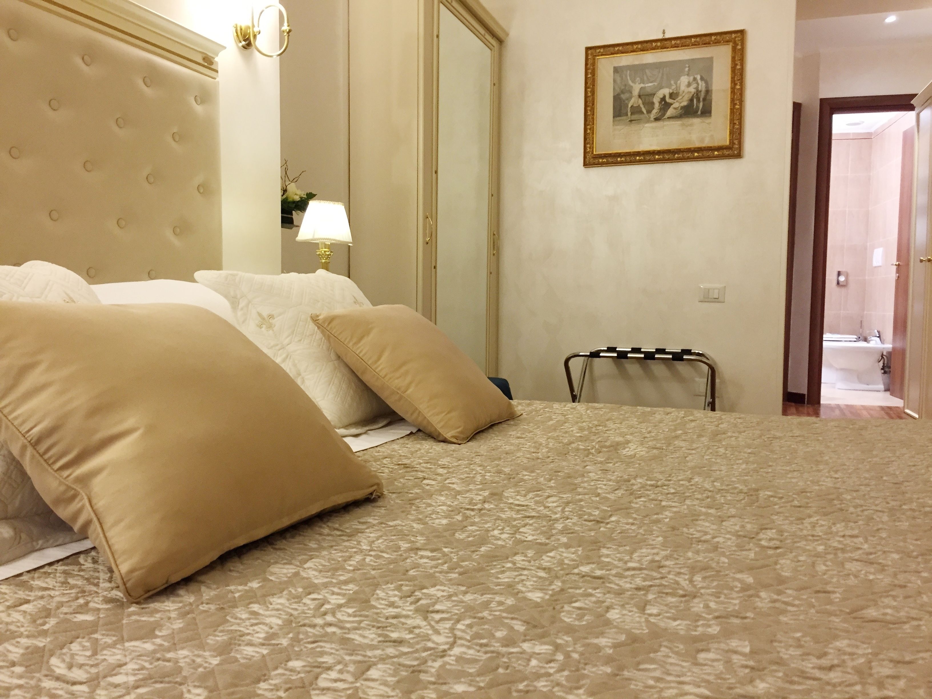A hotel room with a large bed, a headboard, a bedside table with a lamp, a mirror, and a framed picture on the wall.