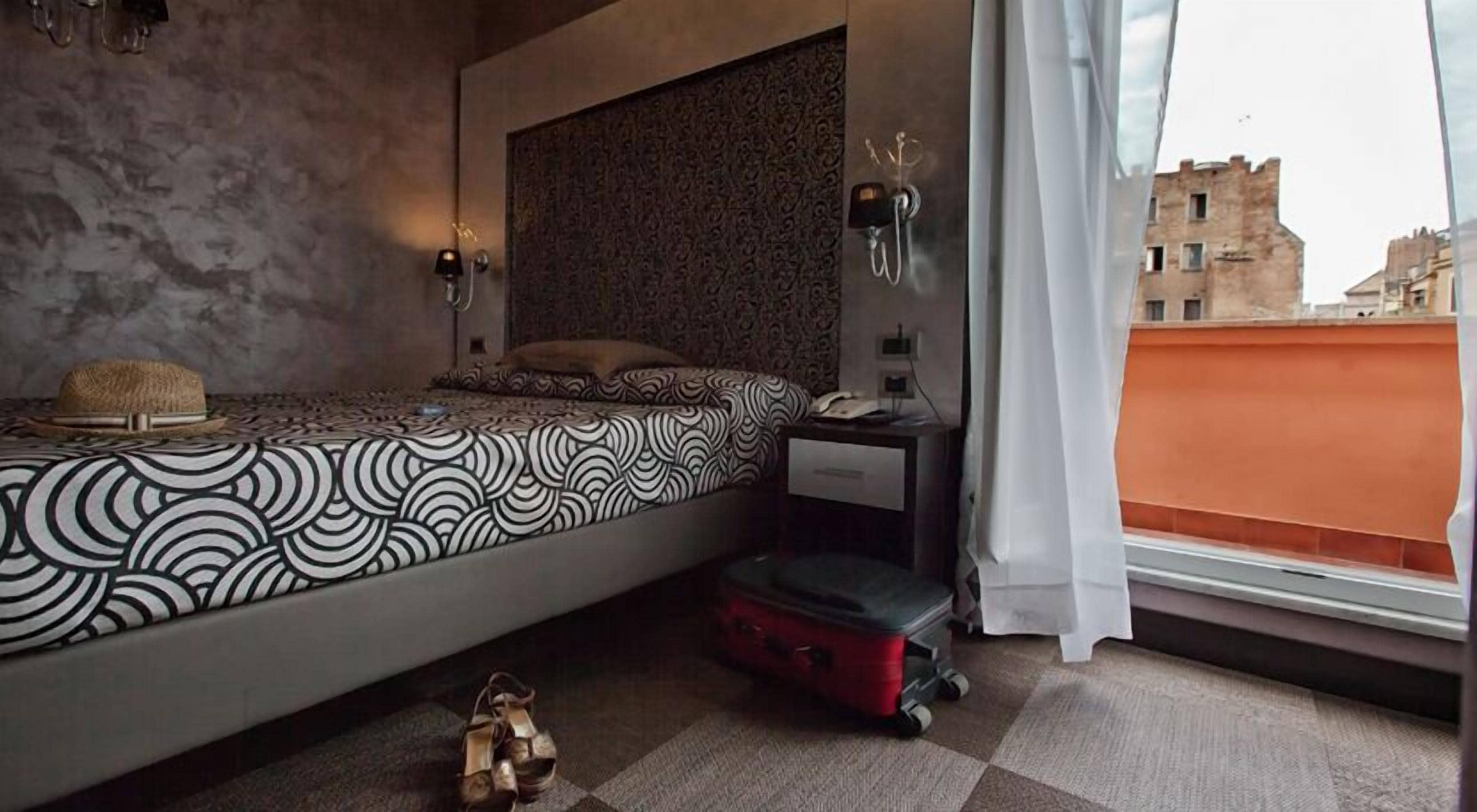 A bedroom with a patterned bedspread, a suitcase, and a city view through a window.