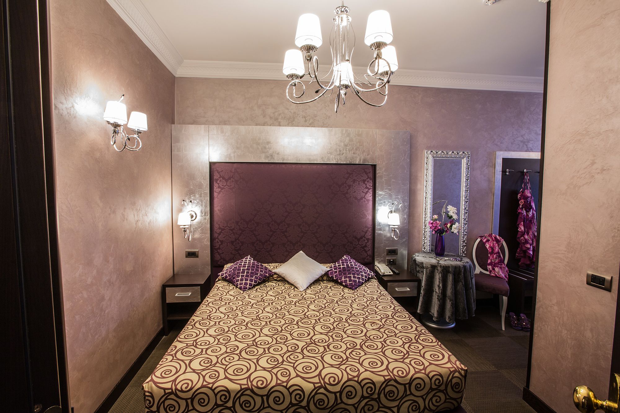 A hotel room with a large bed, a chandelier, a vanity with a mirror, and a robe hanging on a hook.