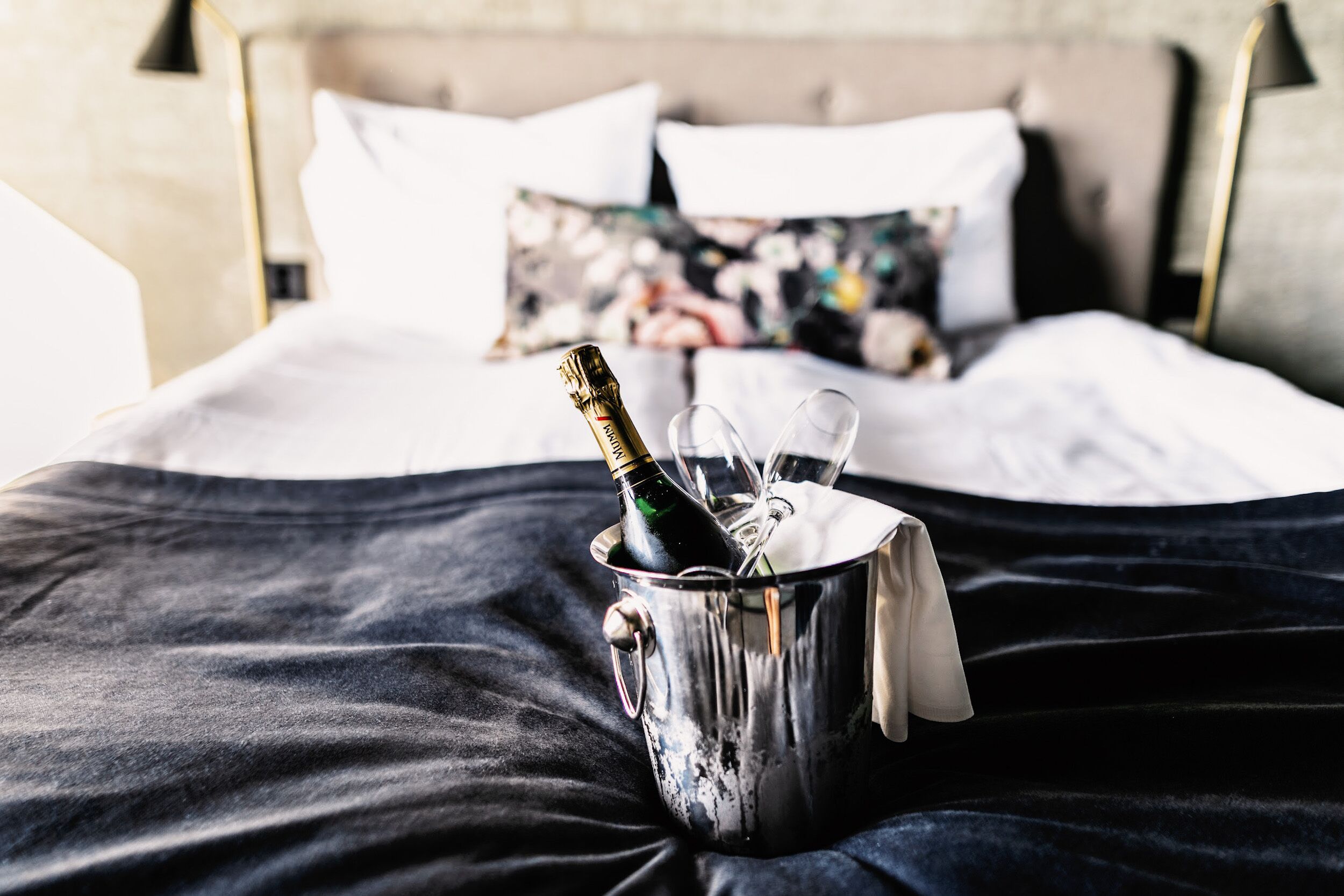 A bed with a metal bucket containing a bottle and glasses, a black bedspread, and a pillow with a floral pattern.