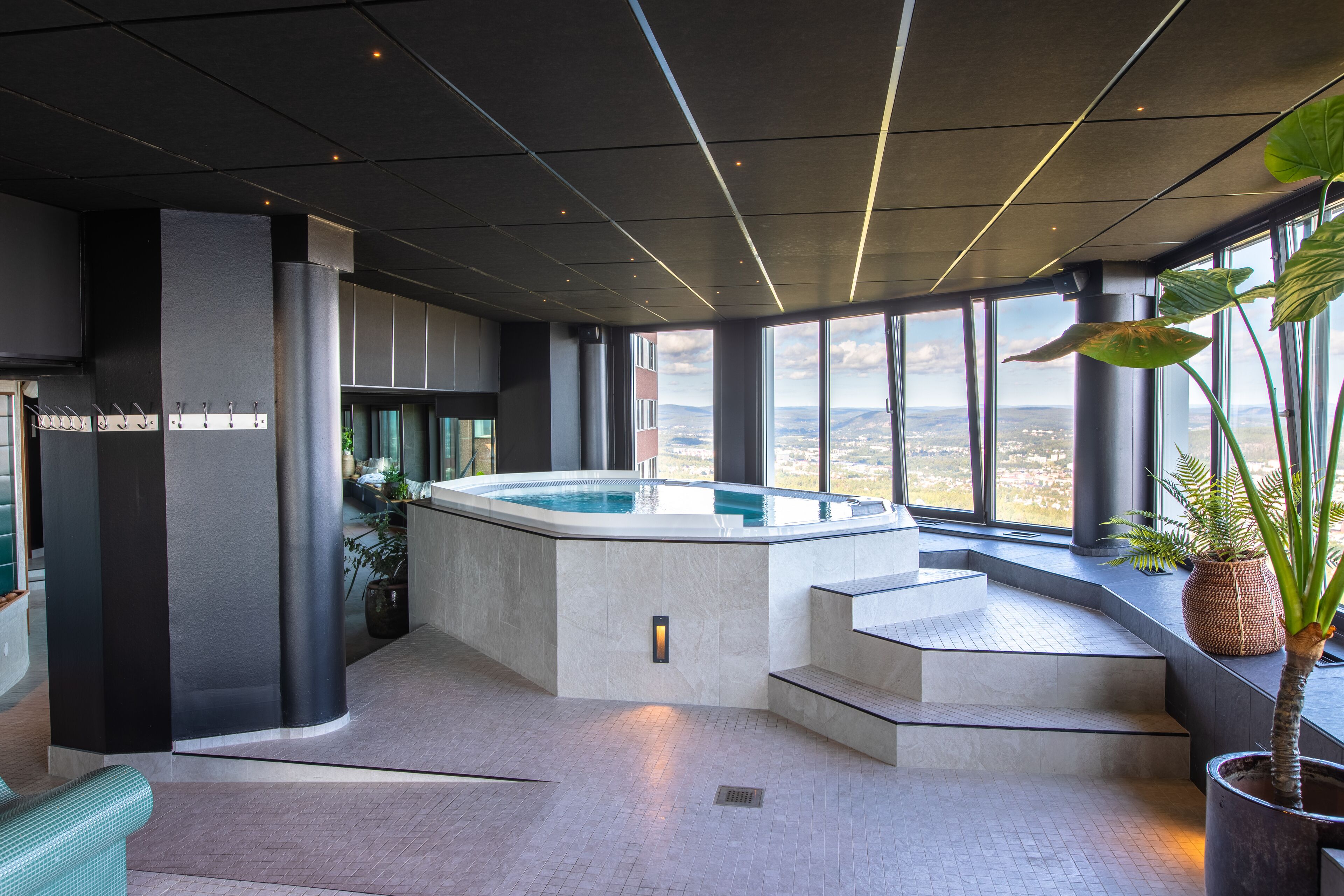 A modern indoor pool area with a view of the cityscape, featuring a large window, a hot tub, and a staircase leading to an upper level.