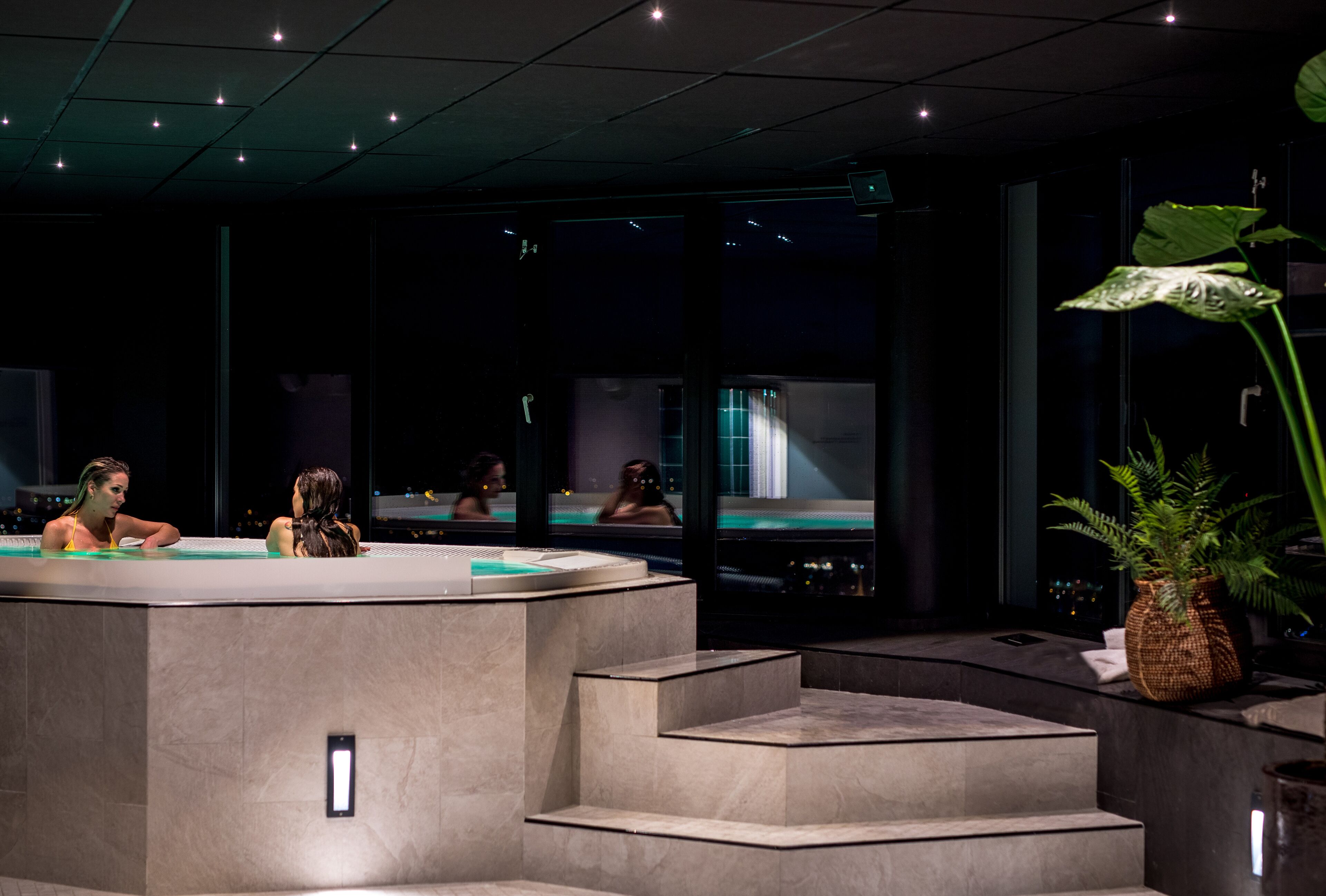 A modern spa area with a reception desk, a seating area, and a large window providing a view of the cityscape at night.