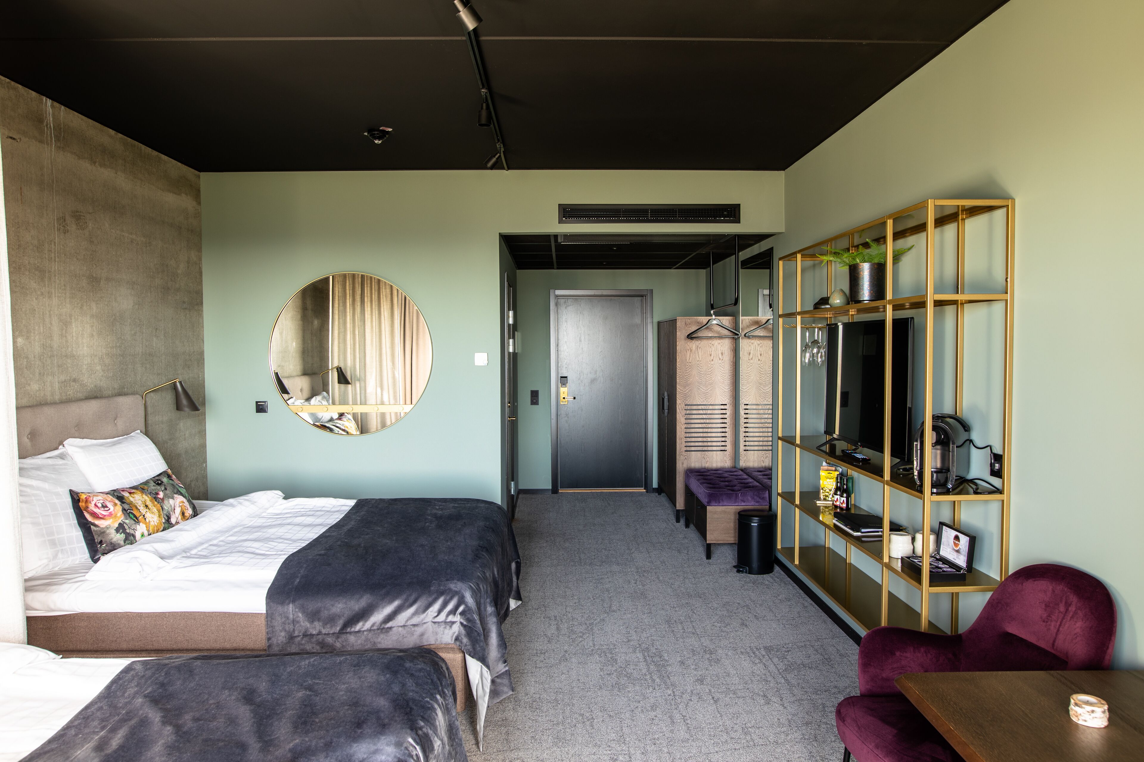 Habitación de hotel con cama, un espejo redondo, una zona de estar con mesa y silla, y una estantería de madera.