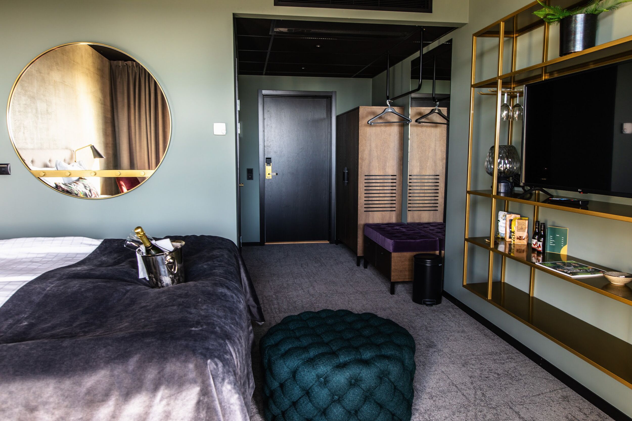 A hotel room with a bed, a round mirror, a wardrobe, a seating area, and a TV on a shelf.